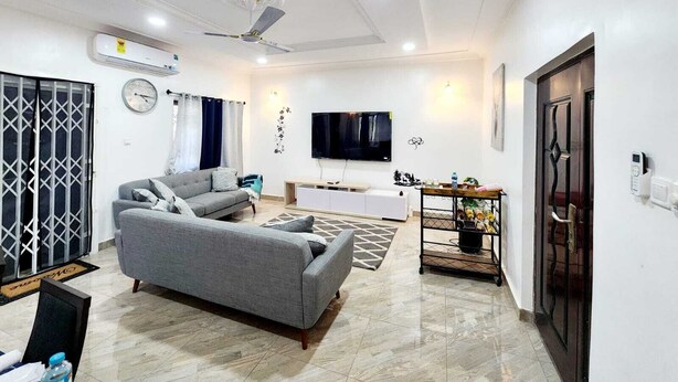 A modern living room with a grey sofa, a TV, a ceiling fan, and a dining area with a table and chairs. - Apartment Vacation Rental Accra Adenta  (MARLEJOR, Adenta Municipality)