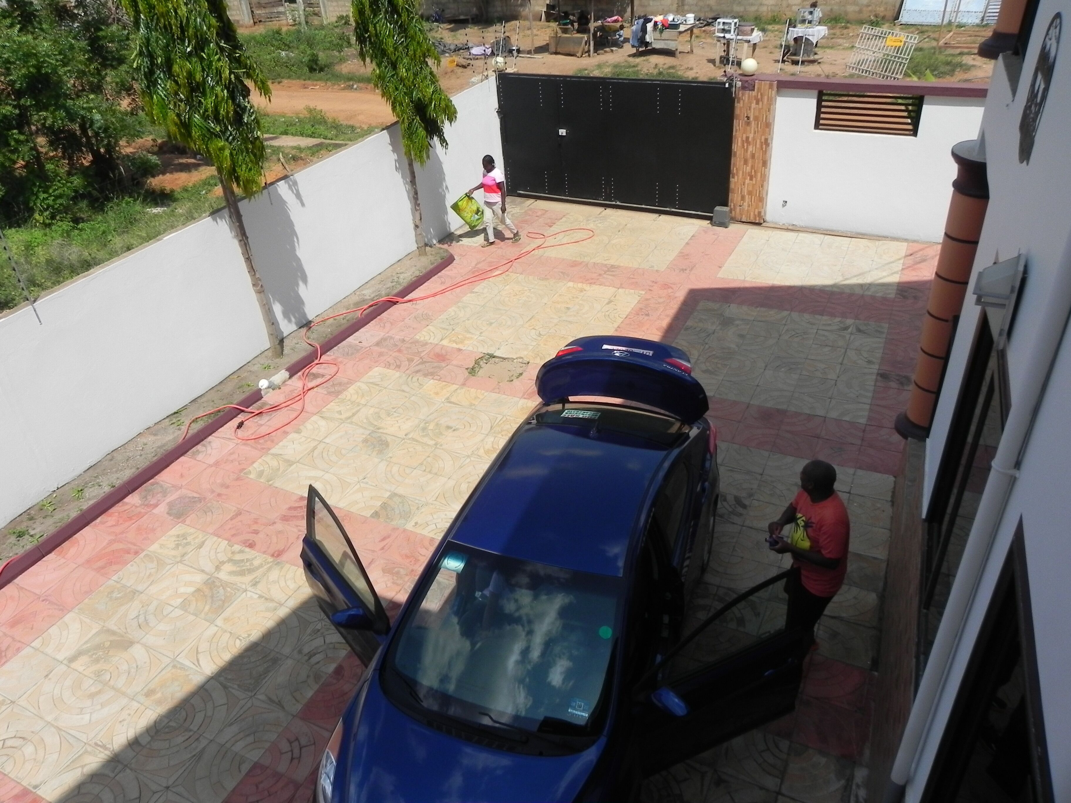 A blue car parked in a covered parking area with a tiled floor, a gated entrance, and a white wall.