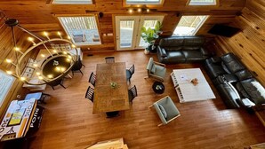 Interior acolhedor de uma cabana com piso de madeira, uma mesa de jantar central, um sofá e uma janela grande.