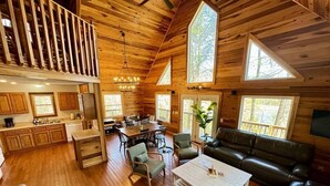 Interior espaçoso de cabana com pisos de madeira, área de jantar, sala de estar com sofá e cadeiras, e cozinha com armários de madeira.