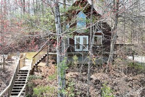 Une cabane avec un escalier en bois menant à un porche, entourée d’arbres et bordée d’une zone boisée.