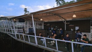A covered outdoor seating area with tables and chairs, a bar with shelves of bottles, and a view of the surrounding landscape. - KEMA Merbabu powered by Cocotel (Ampel)
