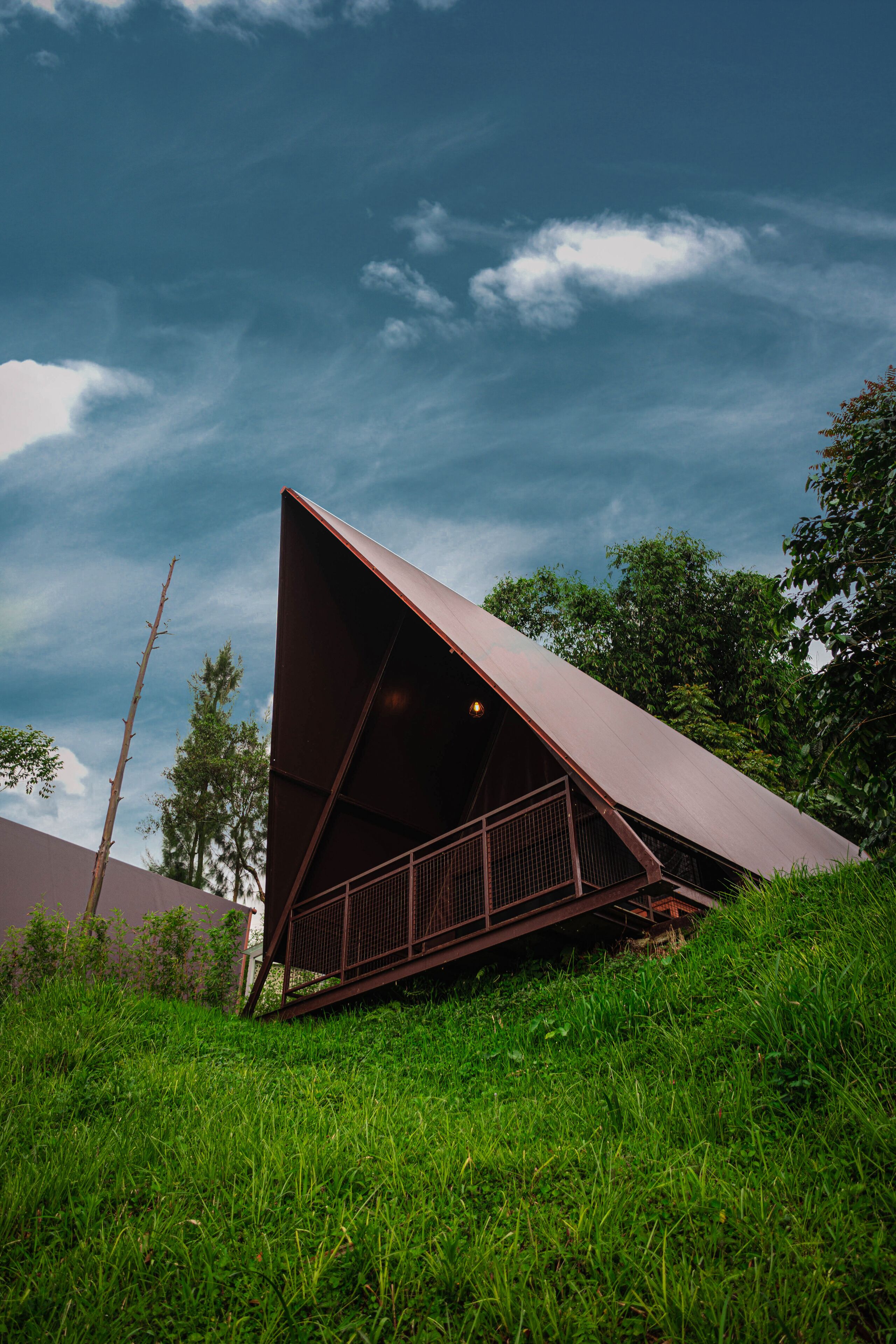 Una estructura metálica triangular moderna con techo, rodeada de abundante hierba verde y árboles, bajo un cielo parcialmente nublado.