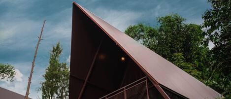 Una estructura metálica triangular moderna con techo, rodeada de abundante hierba verde y árboles, bajo un cielo parcialmente nublado.