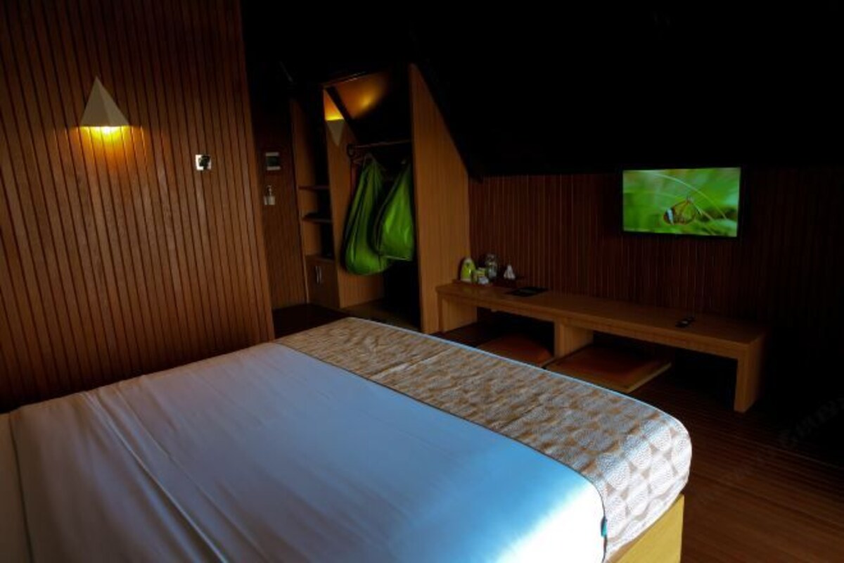A room with a bed, a wooden bench, a television, and a green bag hanging on the wall.