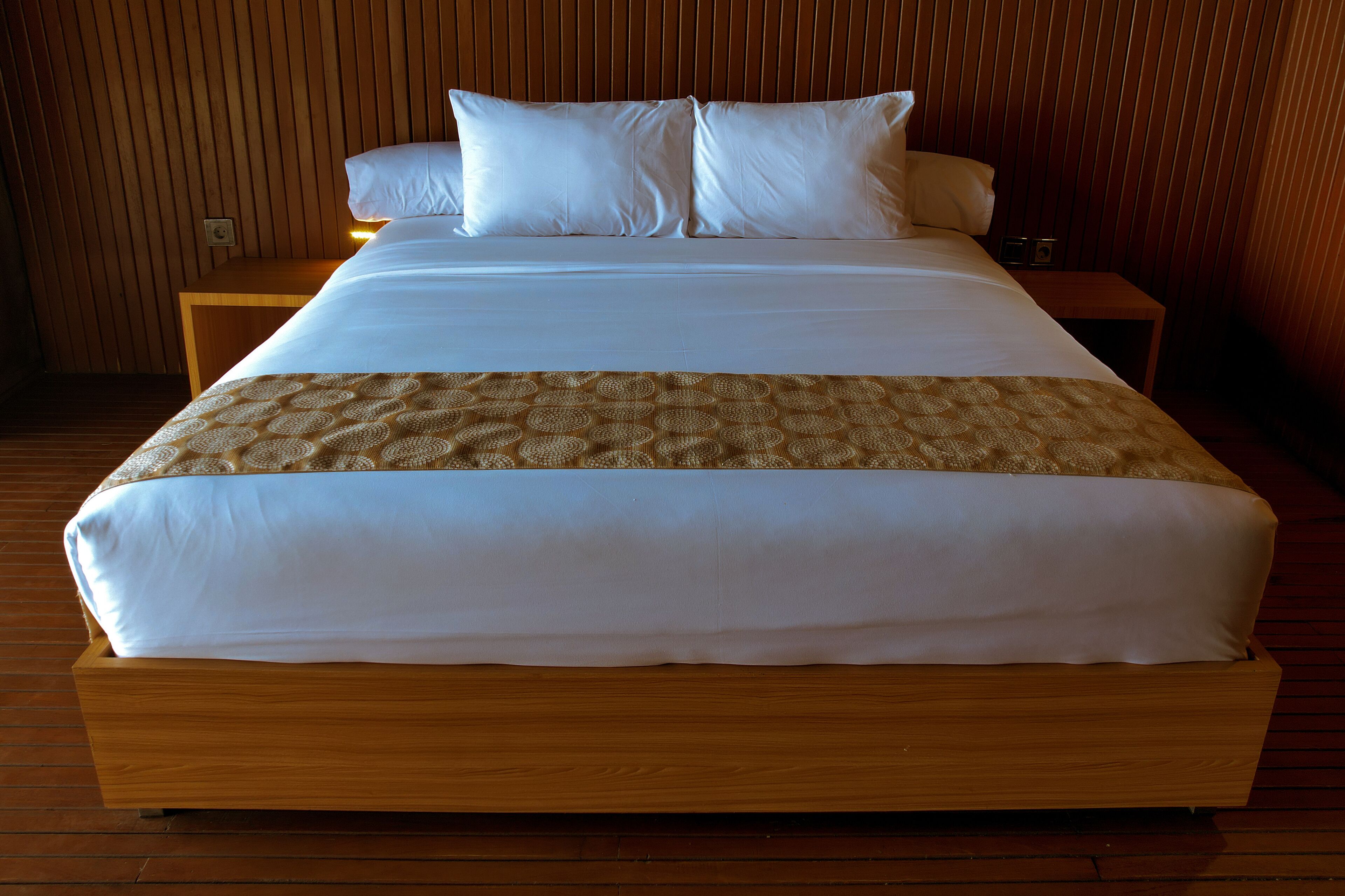 Ein großes Bett mit weißer Bettwäsche und einer gemusterten Tagesdecke, vor einer Holzwand mit einer Sparrenstruktur.