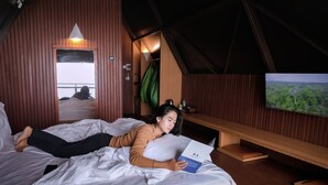 A room with a bed, a wooden bench, a television, and a view of a forest. - KEMA Merbabu powered by Cocotel (Ampel)