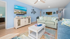 Un salon doté d’un ventilateur de plafond, d’un téléviseur fixé au mur, d’un canapé de couleur claire, d’un canapé bleu et d’une table basse sur laquelle se trouve un bol décoratif.