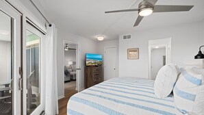 A bedroom with a large bed, a ceiling fan, a TV, and a view of a beach through a sliding door.