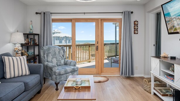 Ein Wohnzimmer mit Sofa, Sessel und Couchtisch, einem Fernseher sowie einem Balkon mit Blick auf Gebäude und das Meer.