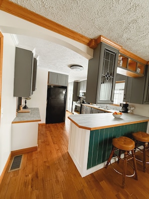 Eine Küche mit Holzboden, einer zentralen Kochinsel mit Hockern, grauen Schränken und einem schwarzen Kühlschrank.