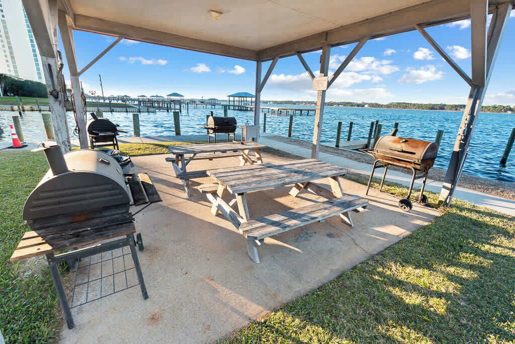 An outdoor covered patio with a grill, picnic table, and seating area overlooking a body of water.