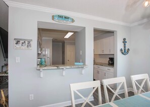 Un comedor con una cocina al fondo, un letrero sobre la ventana que dice "THIS WAY to the beach", y una ancla decorativa en la pared.