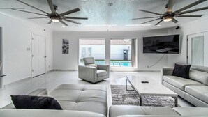 A spacious living room with a large TV, two ceiling fans, a pool view, and a modern design. - Ocean View + Poolside Opulence Tennis (Corpus Christi)