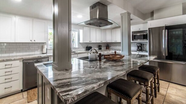 A modern kitchen with white cabinetry, a marble island, stainless steel appliances, and a large window. - Sail Away! Your Coastal Pool Oasis Awaits (Corpus Christi)