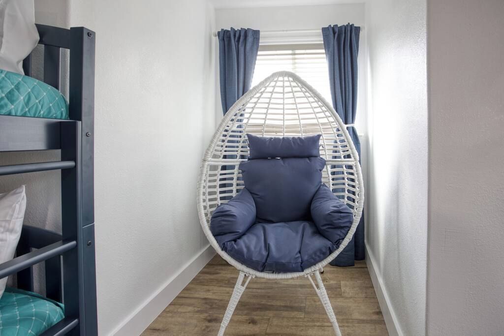 Een smalle gang met een witte rieten stoel met blauwe kussens, een houten stapelbed met blauwgroene en witte beddengoed, en een raam met blauwe gordijnen.