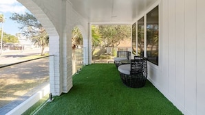 A covered patio with artificial grass, a wicker table and chairs, and a view of the street and trees. - Corpus Christi Gem (Corpus Christi)