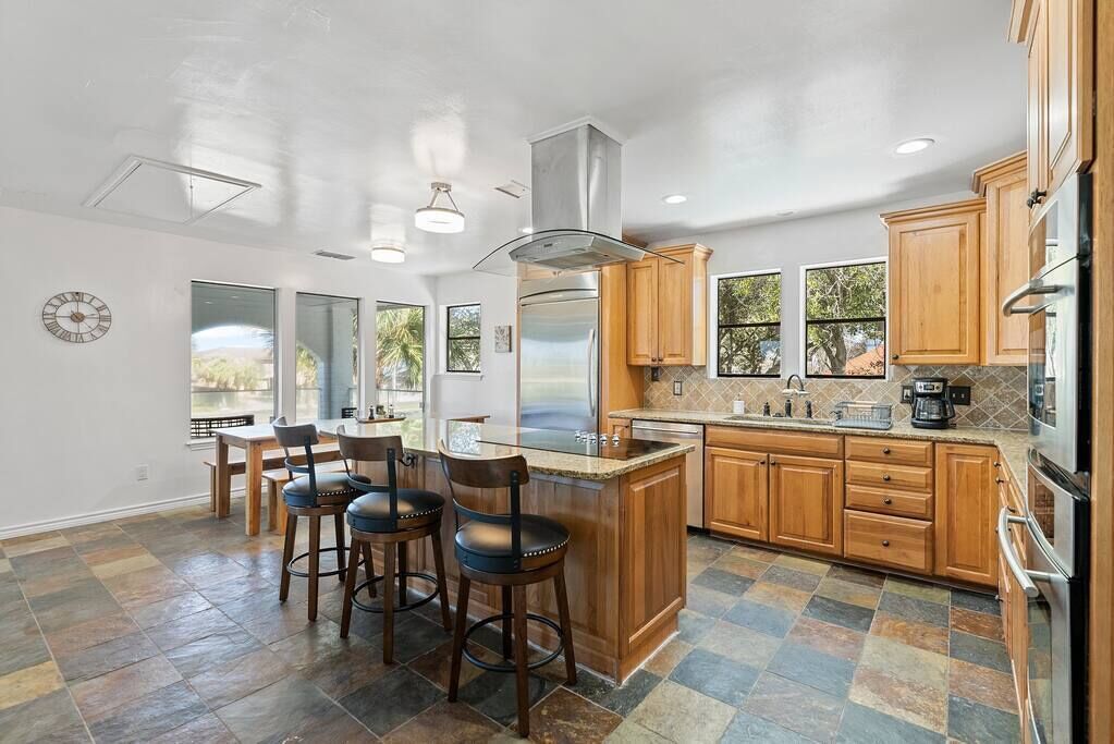 A modern kitchen with wooden cabinets, a central island with stools, a stainless steel refrigerator, and a large window providing natural light.