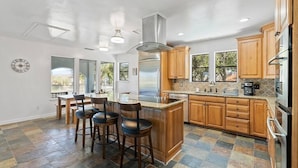 A modern kitchen with wooden cabinets, a central island with stools, a stainless steel refrigerator, and a large window providing natural light. - Corpus Christi Gem (Corpus Christi)