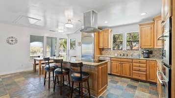 A modern kitchen with wooden cabinets, a central island with stools, a stainless steel refrigerator, and a large window providing natural light.