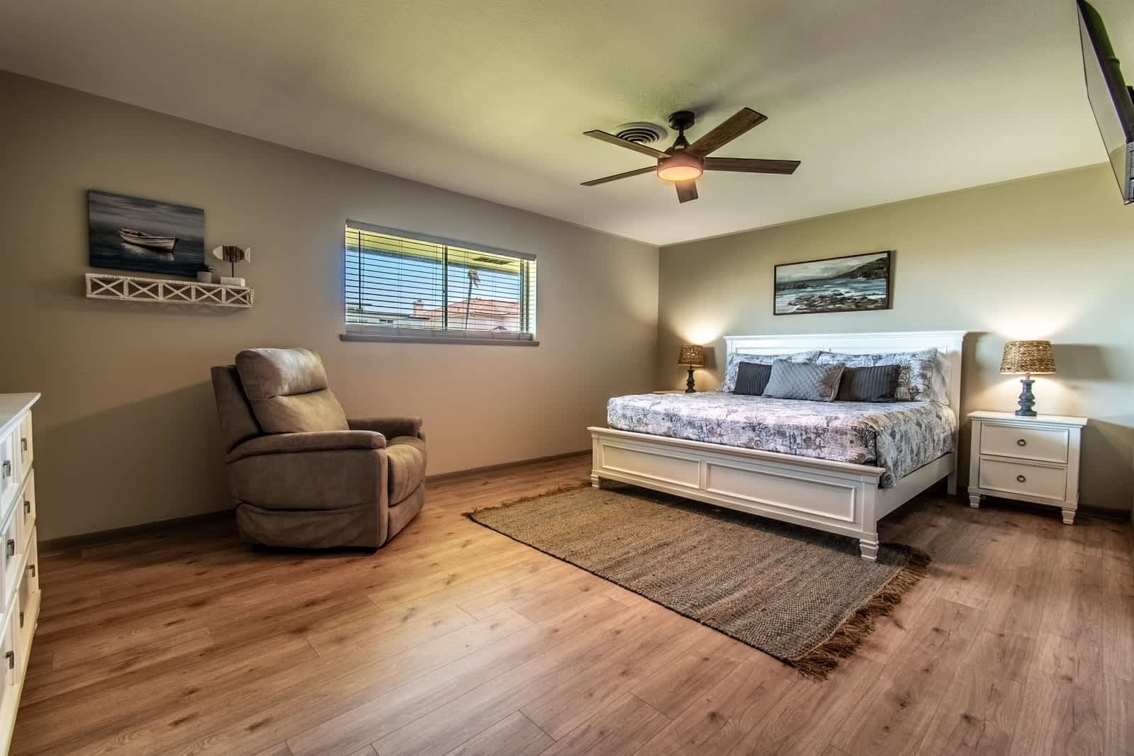 A bedroom with a bed, a chair, a nightstand, a ceiling fan, and two windows.