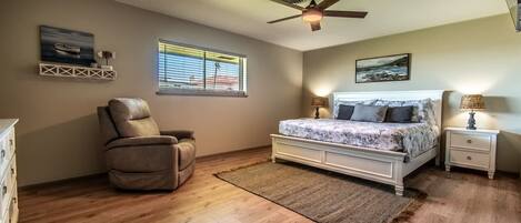 A bedroom with a bed, a chair, a nightstand, a ceiling fan, and two windows.