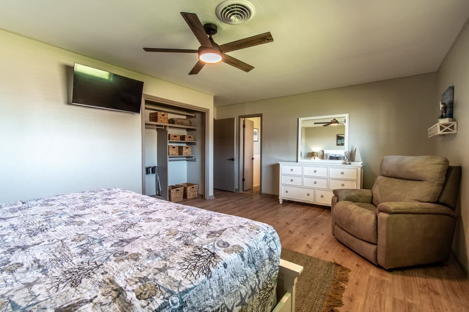 Une chambre à coucher avec un lit, un ventilateur de plafond, une commode, une chaise et un téléviseur fixé au mur.