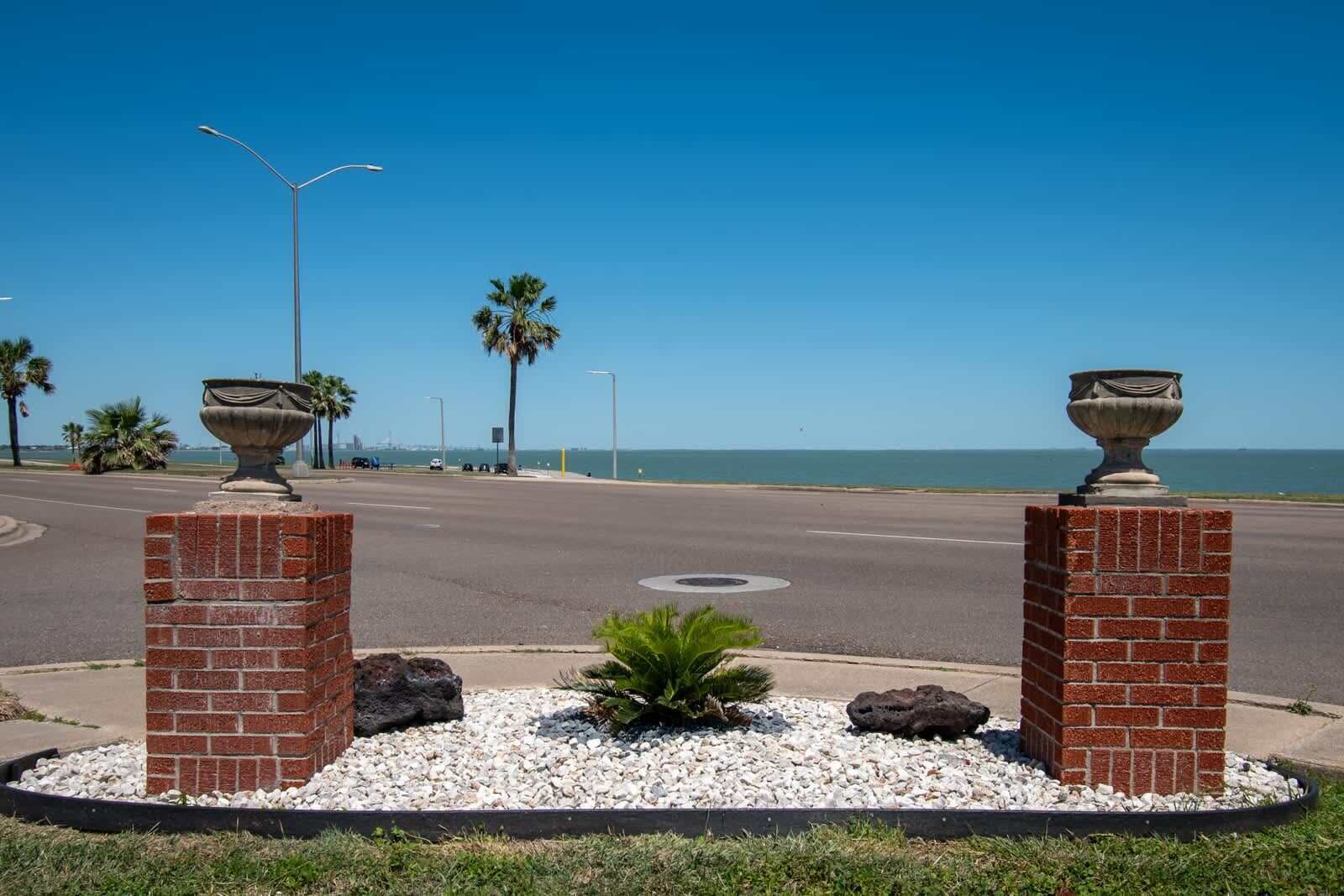 A street view with two brick pillars supporting large urns, a palm tree, and a clear blue sky.