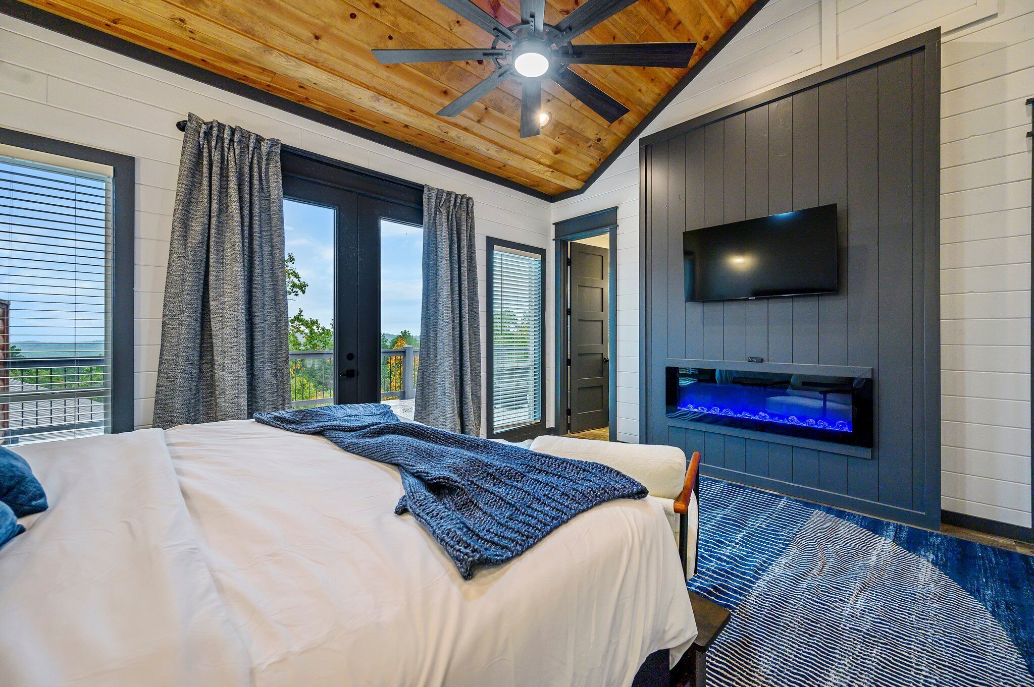 A bedroom with a bed, a ceiling fan, a TV mounted on the wall, and a window with curtains.