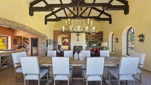 Un comedor amplio con una gran mesa de madera rodeada de sillas blancas con tapicería, una lámpara de araña decorativa de hierro forjado y un techo abovedado con vigas a la vista.