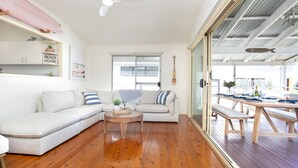 Un salon moderne avec un canapé blanc, une table basse en bois et une porte vitrée donnant sur un espace repas extérieur.