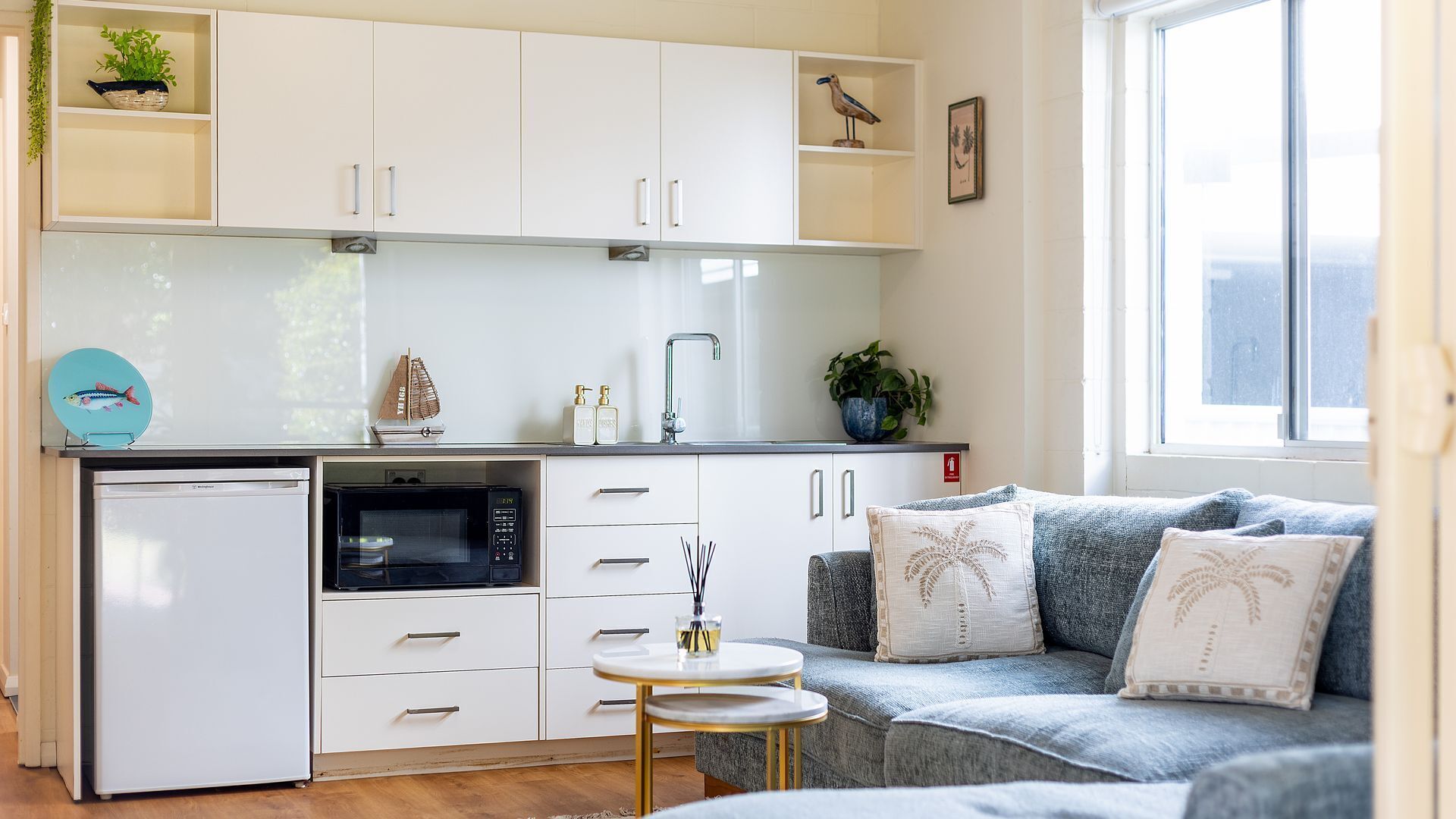 A compact kitchen with white cabinets, a microwave, and a sink. There's a small table with a decorative item and a sofa with patterned cushions.