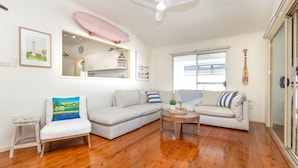 Un salon moderne avec un plancher en bois, un canapé d’angle gris, un petit fauteuil blanc, une table basse et un ventilateur de plafond.