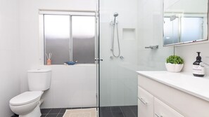 Une salle de bain moderne avec une douche à l’italienne en verre, des toilettes, un lavabo avec un miroir et une petite fenêtre située au-dessus des toilettes.