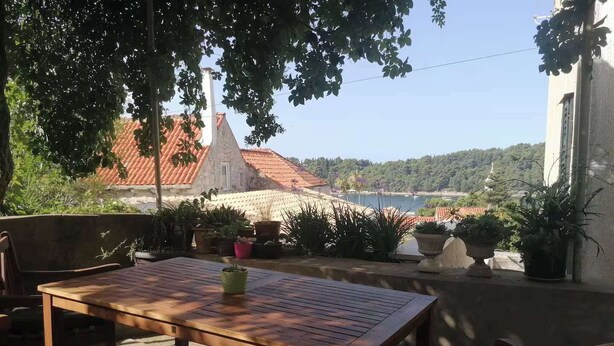 An outdoor patio with a wooden table and chairs, surrounded by potted plants and overlooking a scenic view of a building with a red roof and a body of water in the distance.