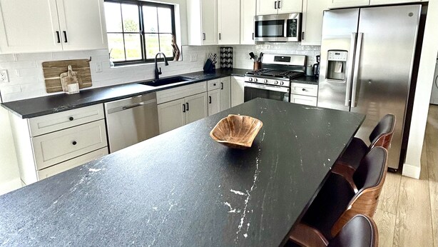 A modern kitchen with white cabinets, a black countertop, stainless steel appliances, and a central island with seating. - Modern Farmhouse w/Privacy, Views and Hot Tub! (GRAND JUNCTION)