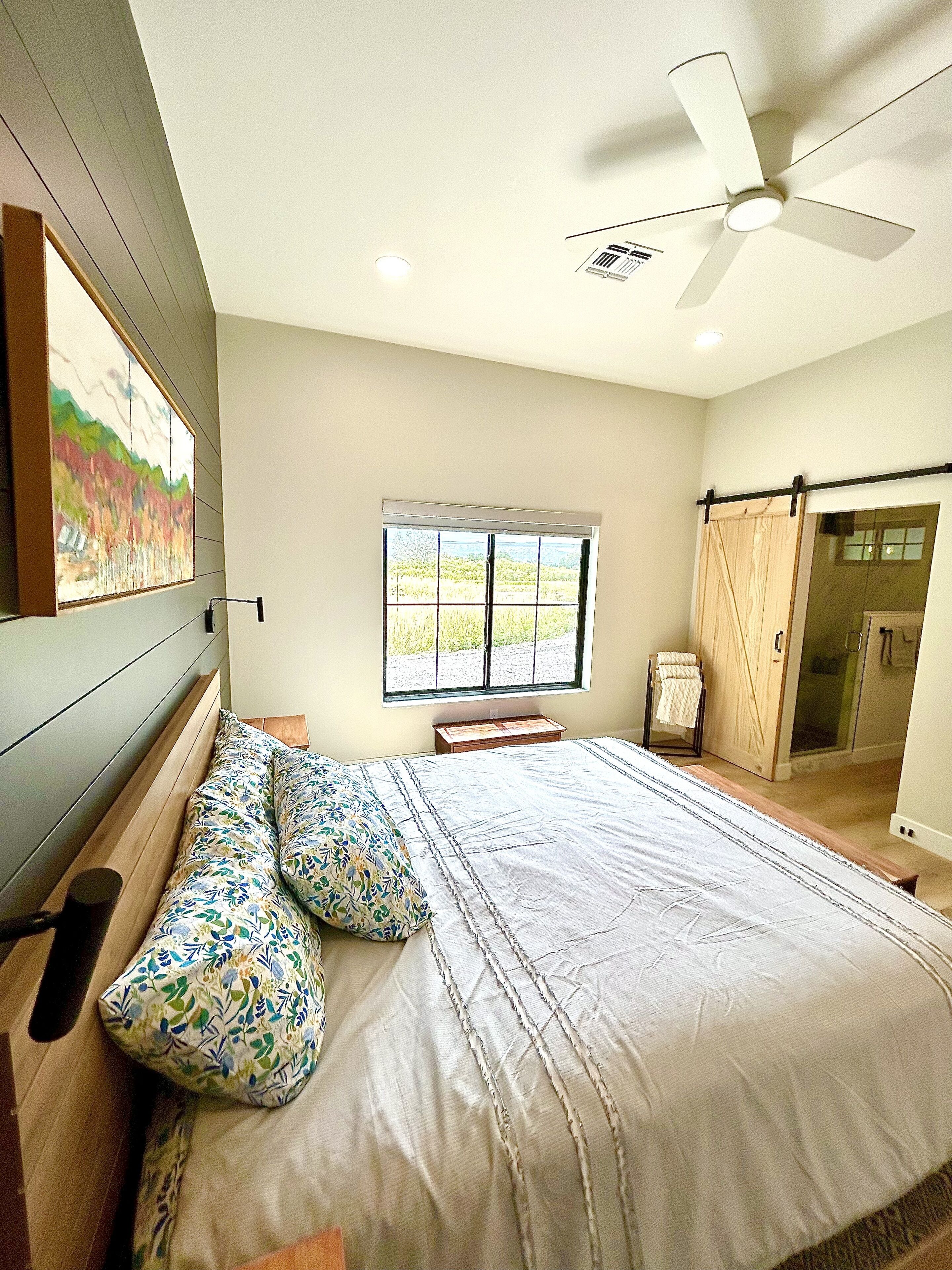 Un dormitorio con una cama, un ventilador de techo, una ventana y una puerta que da a un baño.