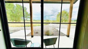 A window with a view of a patio area, including a table and chairs, and a gravel path leading to a grassy field. - Modern Farmhouse w/Privacy, Views and Hot Tub! (GRAND JUNCTION)