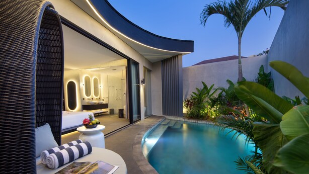A modern outdoor pool area with a curved wall, a lounge chair, and a small table with a fruit bowl. - Je Ne Sais Quoi Seminyak (Seminyak)