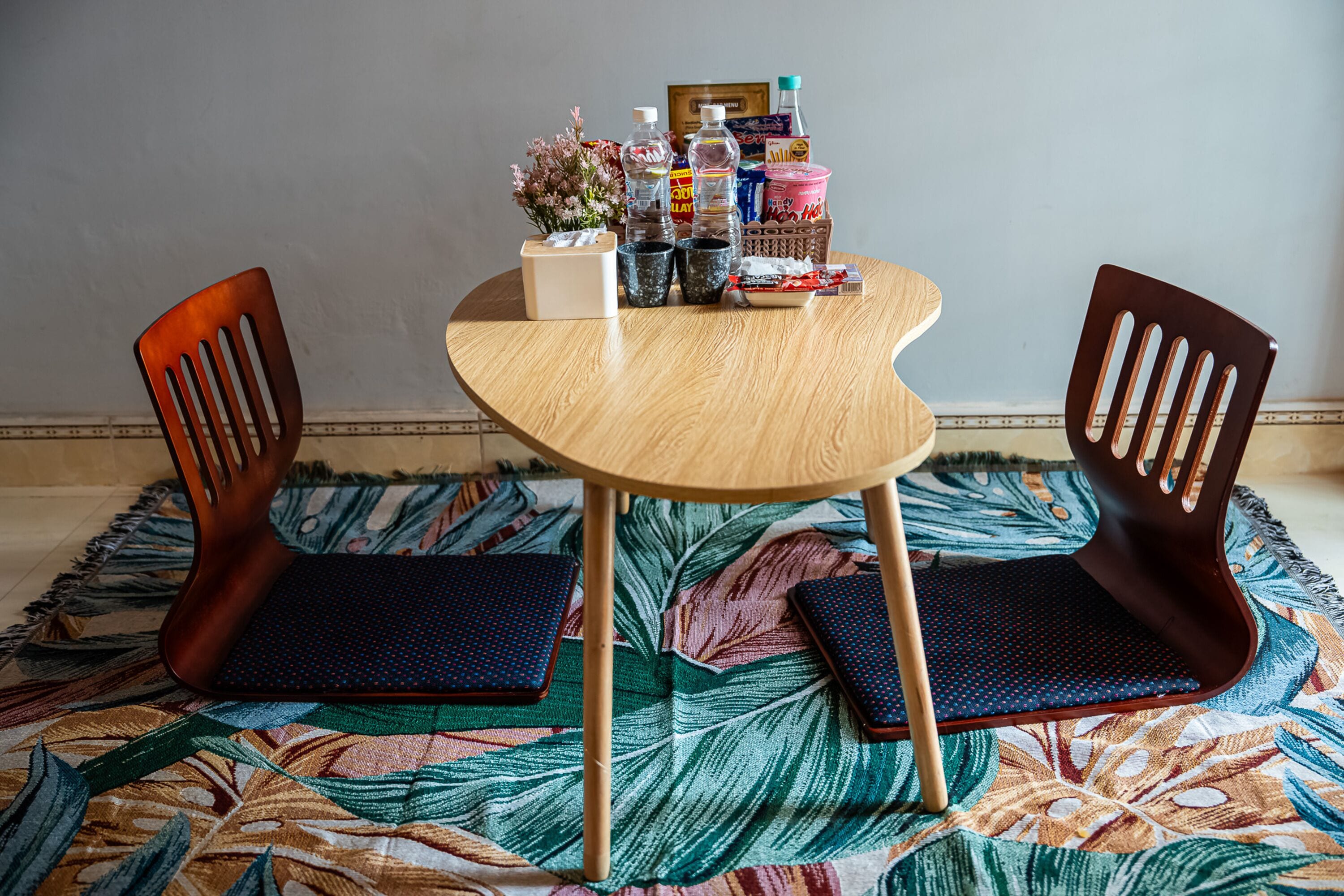 A round wooden table with two chairs, a patterned rug, and various items on the table including a tissue box, a bottle, and snacks.