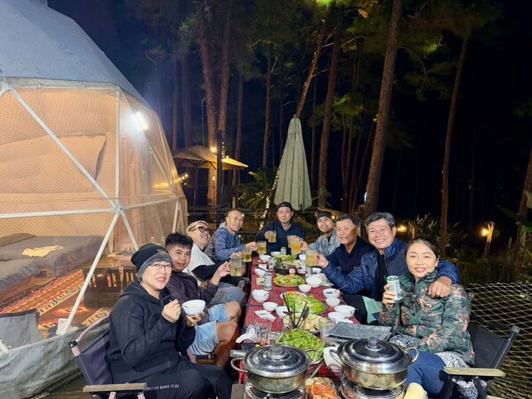 Eine Gruppe von Menschen speist abends im Freien, mit einem großen Zelt und einem Tisch, der mit verschiedenen Speisen und Getränken gedeckt ist.