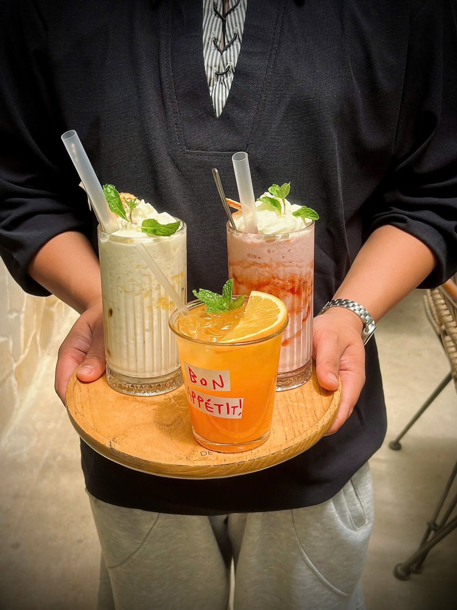Una persona sostiene una bandeja de madera con tres bebidas, cada una con pajita y decorada con hojas de menta y una rodaja de naranja.