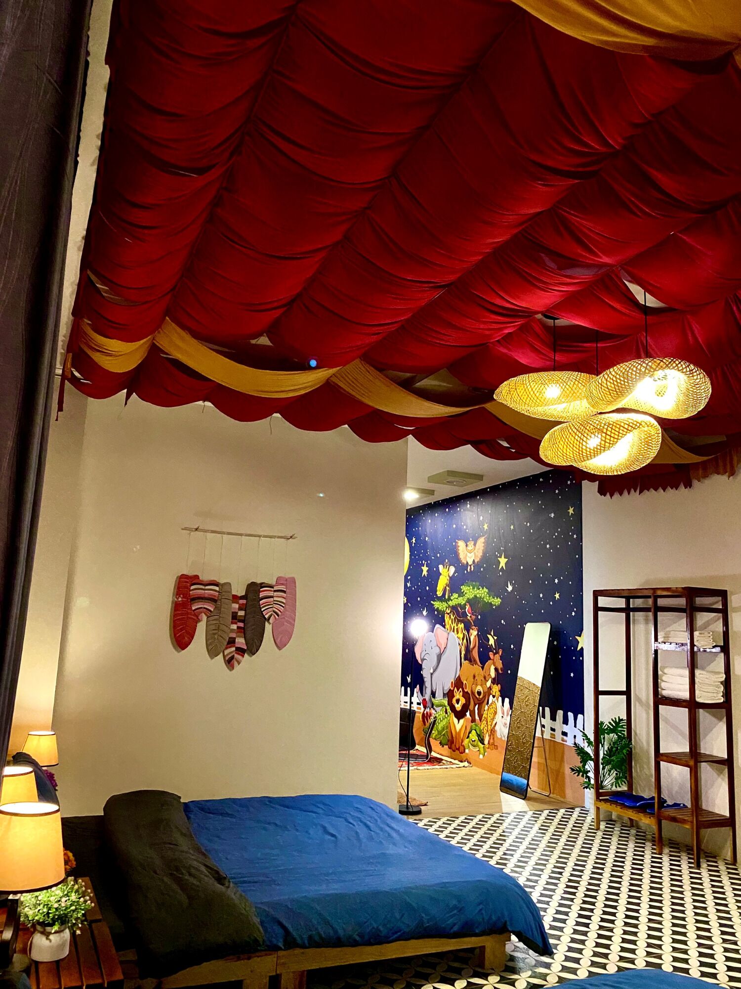 A bedroom with a bed, a hanging lamp, a wall with a night scene mural, and a shelf with towels.