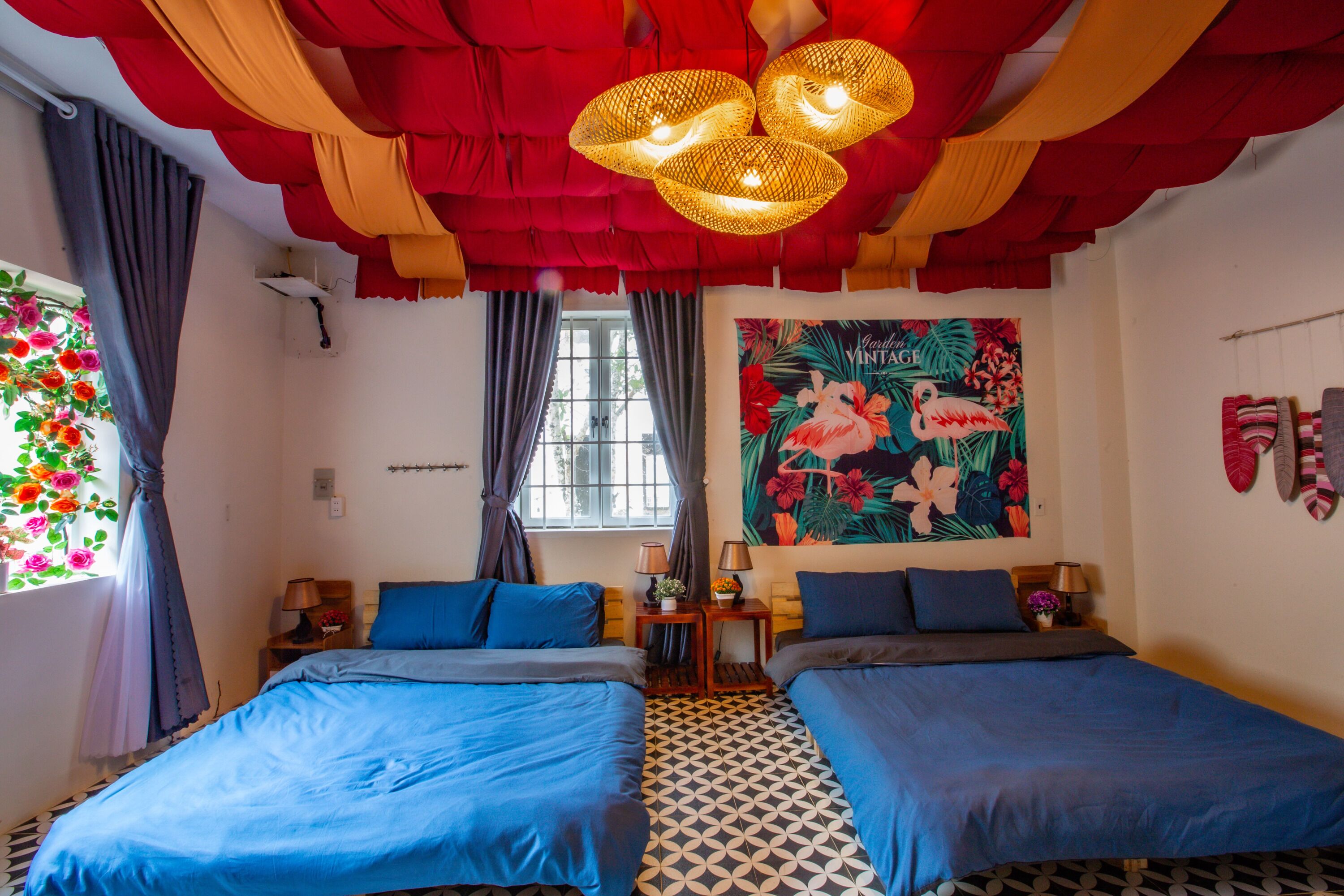 A room with two beds, a window with curtains, a wall with a tropical-themed painting, and a patterned floor.