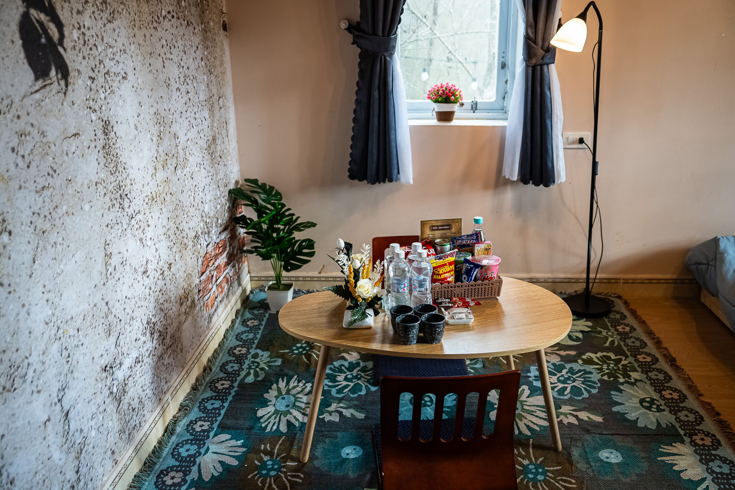 Ein Zimmer mit einem runden Holztisch, Stühlen, einer Topfpflanze, einem kleinen Fenster mit Vorhängen und einer Stehlampe.