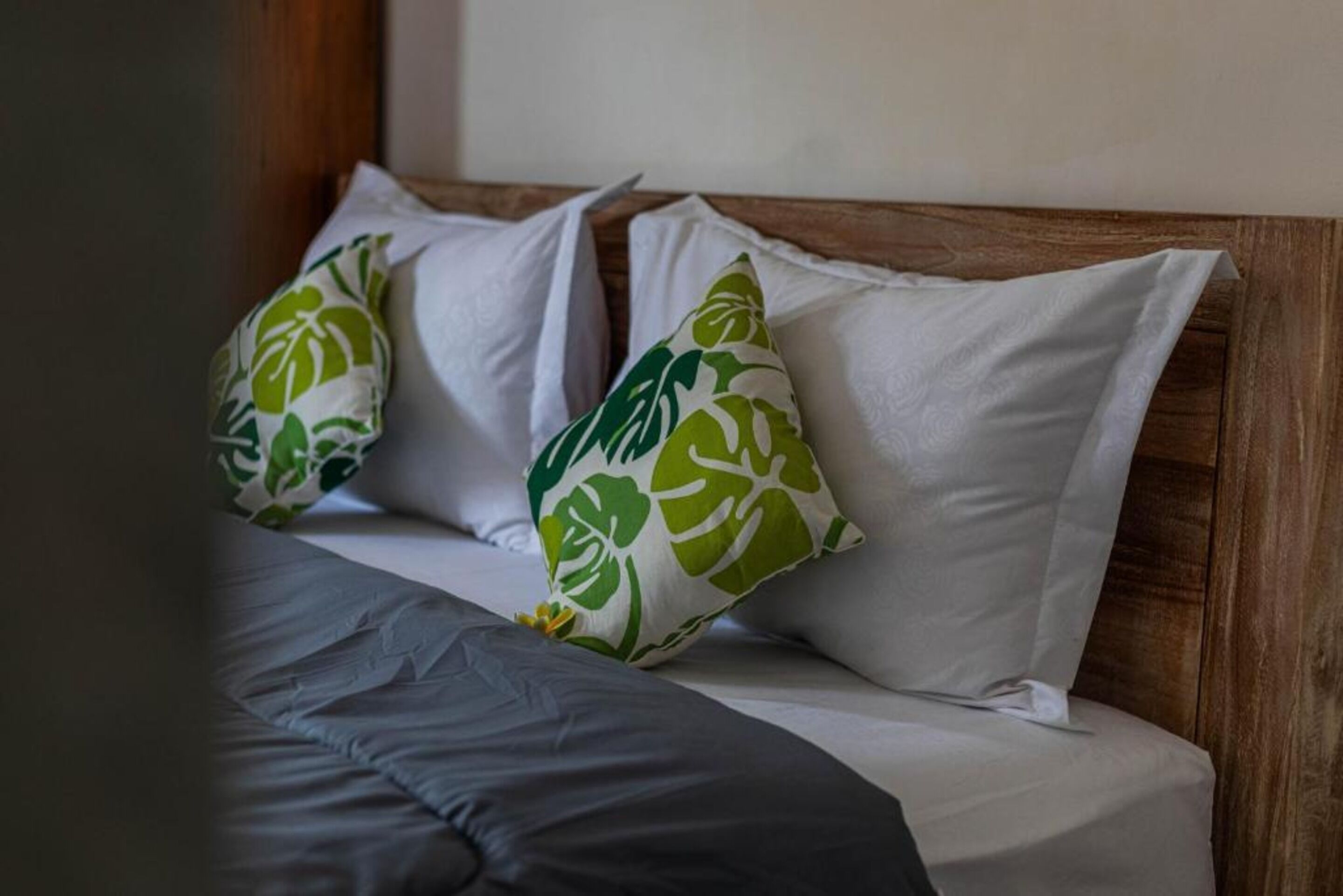 Una cama con almohadas blancas y un estampado de hojas verdes, cabecera de madera y manta gris.
