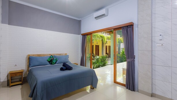 Quarto com cama, uma pequena mesa de cabeceira de madeira, uma porta de vidro deslizante para uma varanda e um ar condicionado branco na parede.