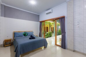 Un dormitorio con cama, una mesita de noche de madera, un ventanal corredizo que da a un balcón y un equipo de aire acondicionado blanco en la pared.