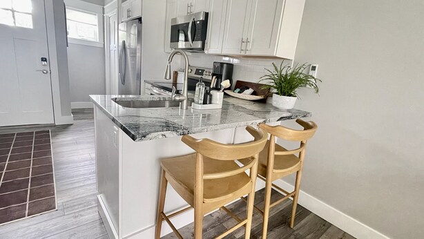 Una cocina moderna con armarios blancos, un refrigerador de acero inoxidable, un microondas y un fregadero. Hay dos sillas de madera en la isla.