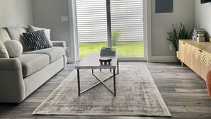 Una sala de estar moderna con un sofá gris, una mesa de centro y un amplio ventanal corredero que da a un espacio exterior verde.